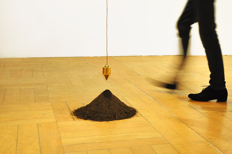 Escultura A Gênese (2011) de Felippe Moraes com prumo banhado a ouro suspenso acima de um montículo de terra