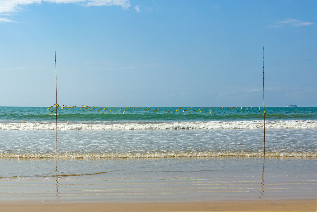 Linha dourada marcando o horizonte na praia enquanto o artista se curva para passar por baixo na obra Aurum'zonte de Felippe Moraes.