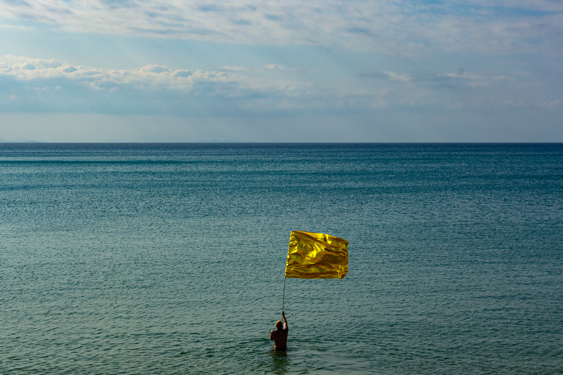 Aurum'zonte, obra de Felippe Moraes da série Prai'Áurea. Na fotografia, uma linha marcada por pequenos fragmentos dourados acompanha o horizonte da praia. O artista se curva para atravessar esse limite imaginário, evocando uma relação lúdica e filosófica com a paisagem e sugerindo a possibilidade de atravessar um limiar simbólico entre corpo, espaço e horizonte.