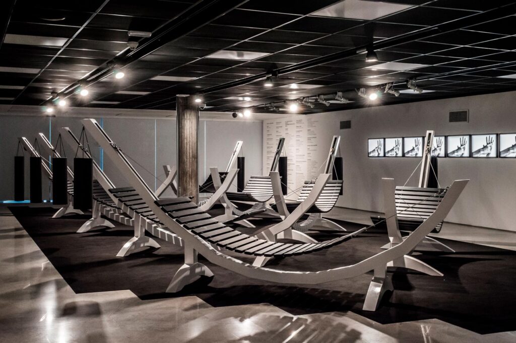 Série Composição Aleatória, 2019 metal escovado, madeira, chapa de aço pintada e cabo de aço 165 x 395 x 96 cm A obra “Composição Aleatória” (2019) foi concebida, construída e instalada pela primeira vez nesta exposição. Oito redes – correspondentes às sete notas musicais acrescidas do Dó da oitava acima – estão presas a tubos sonoros e badalos que vibram e ecoam dependendo dos movimentos a que são submetidos. Nossos corpos são novamente convidados a participar da criação do trabalho, ativando os balanços e ressoando os sinos. A composição cresce e se complexifica à medida que mais pessoas juntam-se à instalação, até que todos os sons sejam entoados em harmonia ou dissonância. Em momentos distintos, composições diferentes preenchem o espaço expositivo – os arranjos dependem, sempre, dos arbitrários e aleatórios gestos dos visitantes, que podem embalar-se com intensidade ou apenas recostar-se em meio à melodia improvisada.