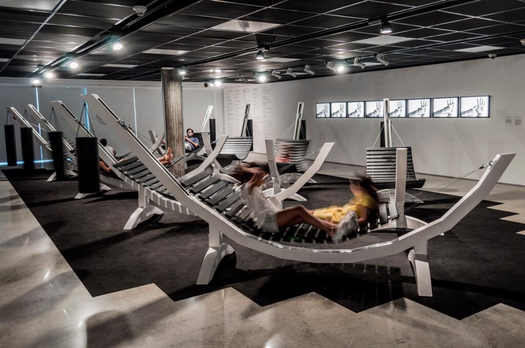 Série Composição Aleatória, 2019 metal escovado, madeira, chapa de aço pintada e cabo de aço 165 x 395 x 96 cm A obra “Composição Aleatória” (2019) foi concebida, construída e instalada pela primeira vez nesta exposição. Oito redes – correspondentes às sete notas musicais acrescidas do Dó da oitava acima – estão presas a tubos sonoros e badalos que vibram e ecoam dependendo dos movimentos a que são submetidos. Nossos corpos são novamente convidados a participar da criação do trabalho, ativando os balanços e ressoando os sinos. A composição cresce e se complexifica à medida que mais pessoas juntam-se à instalação, até que todos os sons sejam entoados em harmonia ou dissonância. Em momentos distintos, composições diferentes preenchem o espaço expositivo – os arranjos dependem, sempre, dos arbitrários e aleatórios gestos dos visitantes, que podem embalar-se com intensidade ou apenas recostar-se em meio à melodia improvisada.