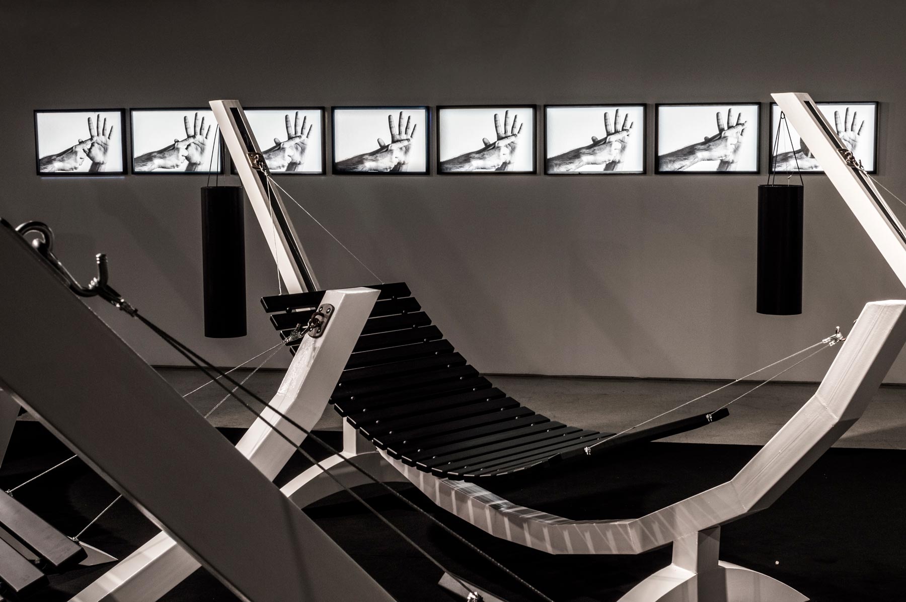 Série Composição Aleatória, 2019 metal escovado, madeira, chapa de aço pintada e cabo de aço 165 x 395 x 96 cm A obra “Composição Aleatória” (2019) foi concebida, construída e instalada pela primeira vez nesta exposição. Oito redes – correspondentes às sete notas musicais acrescidas do Dó da oitava acima – estão presas a tubos sonoros e badalos que vibram e ecoam dependendo dos movimentos a que são submetidos. Nossos corpos são novamente convidados a participar da criação do trabalho, ativando os balanços e ressoando os sinos. A composição cresce e se complexifica à medida que mais pessoas juntam-se à instalação, até que todos os sons sejam entoados em harmonia ou dissonância. Em momentos distintos, composições diferentes preenchem o espaço expositivo – os arranjos dependem, sempre, dos arbitrários e aleatórios gestos dos visitantes, que podem embalar-se com intensidade ou apenas recostar-se em meio à melodia improvisada.