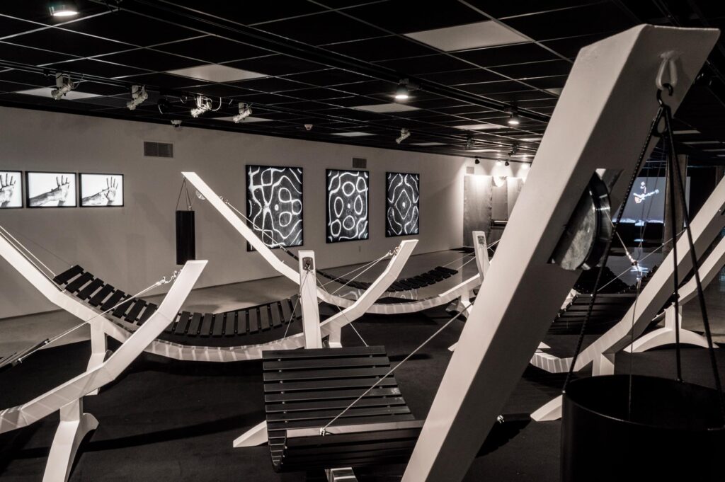 Série Composição Aleatória, 2019 metal escovado, madeira, chapa de aço pintada e cabo de aço 165 x 395 x 96 cm A obra “Composição Aleatória” (2019) foi concebida, construída e instalada pela primeira vez nesta exposição. Oito redes – correspondentes às sete notas musicais acrescidas do Dó da oitava acima – estão presas a tubos sonoros e badalos que vibram e ecoam dependendo dos movimentos a que são submetidos. Nossos corpos são novamente convidados a participar da criação do trabalho, ativando os balanços e ressoando os sinos. A composição cresce e se complexifica à medida que mais pessoas juntam-se à instalação, até que todos os sons sejam entoados em harmonia ou dissonância. Em momentos distintos, composições diferentes preenchem o espaço expositivo – os arranjos dependem, sempre, dos arbitrários e aleatórios gestos dos visitantes, que podem embalar-se com intensidade ou apenas recostar-se em meio à melodia improvisada.