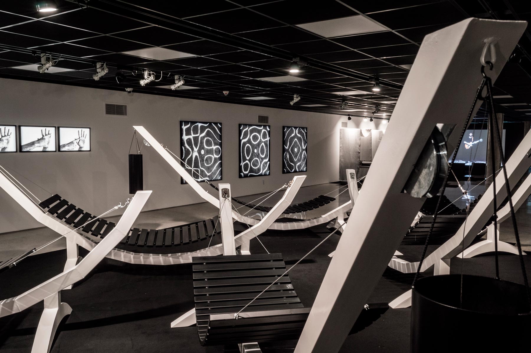 Série Composição Aleatória, 2019 metal escovado, madeira, chapa de aço pintada e cabo de aço 165 x 395 x 96 cm A obra “Composição Aleatória” (2019) foi concebida, construída e instalada pela primeira vez nesta exposição. Oito redes – correspondentes às sete notas musicais acrescidas do Dó da oitava acima – estão presas a tubos sonoros e badalos que vibram e ecoam dependendo dos movimentos a que são submetidos. Nossos corpos são novamente convidados a participar da criação do trabalho, ativando os balanços e ressoando os sinos. A composição cresce e se complexifica à medida que mais pessoas juntam-se à instalação, até que todos os sons sejam entoados em harmonia ou dissonância. Em momentos distintos, composições diferentes preenchem o espaço expositivo – os arranjos dependem, sempre, dos arbitrários e aleatórios gestos dos visitantes, que podem embalar-se com intensidade ou apenas recostar-se em meio à melodia improvisada.