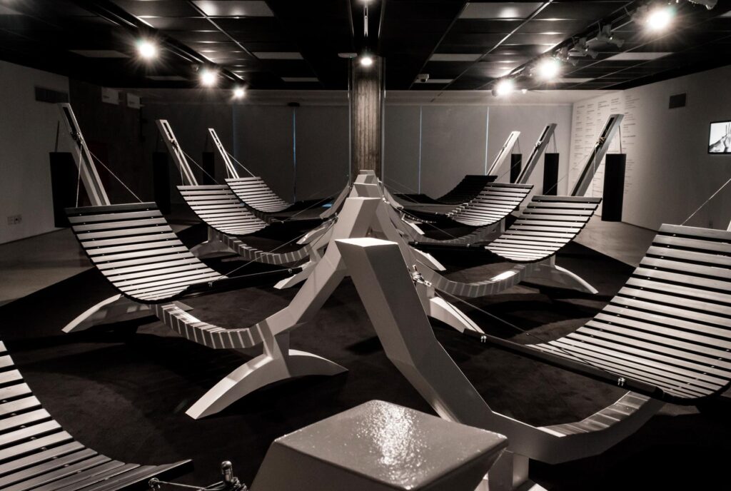 Série Composição Aleatória, 2019 metal escovado, madeira, chapa de aço pintada e cabo de aço 165 x 395 x 96 cm A obra “Composição Aleatória” (2019) foi concebida, construída e instalada pela primeira vez nesta exposição. Oito redes – correspondentes às sete notas musicais acrescidas do Dó da oitava acima – estão presas a tubos sonoros e badalos que vibram e ecoam dependendo dos movimentos a que são submetidos. Nossos corpos são novamente convidados a participar da criação do trabalho, ativando os balanços e ressoando os sinos. A composição cresce e se complexifica à medida que mais pessoas juntam-se à instalação, até que todos os sons sejam entoados em harmonia ou dissonância. Em momentos distintos, composições diferentes preenchem o espaço expositivo – os arranjos dependem, sempre, dos arbitrários e aleatórios gestos dos visitantes, que podem embalar-se com intensidade ou apenas recostar-se em meio à melodia improvisada.