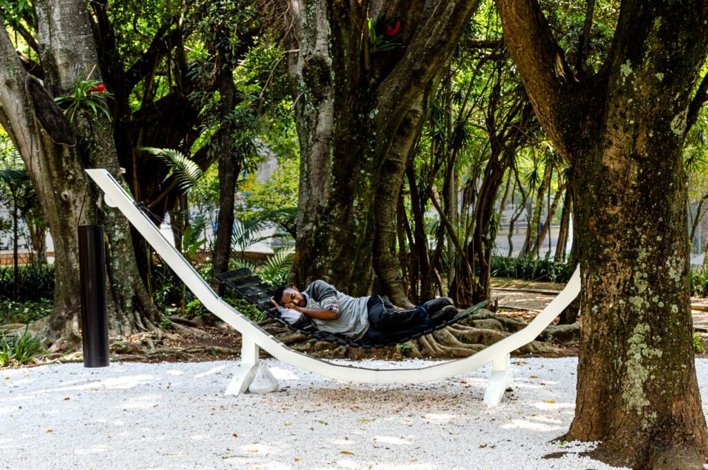 Série Composição Aleatória, 2019 metal escovado, madeira, chapa de aço pintada e cabo de aço 165 x 395 x 96 cm A obra “Composição Aleatória” (2019) foi concebida, construída e instalada pela primeira vez nesta exposição. Oito redes – correspondentes às sete notas musicais acrescidas do Dó da oitava acima – estão presas a tubos sonoros e badalos que vibram e ecoam dependendo dos movimentos a que são submetidos. Nossos corpos são novamente convidados a participar da criação do trabalho, ativando os balanços e ressoando os sinos. A composição cresce e se complexifica à medida que mais pessoas juntam-se à instalação, até que todos os sons sejam entoados em harmonia ou dissonância. Em momentos distintos, composições diferentes preenchem o espaço expositivo – os arranjos dependem, sempre, dos arbitrários e aleatórios gestos dos visitantes, que podem embalar-se com intensidade ou apenas recostar-se em meio à melodia improvisada.
