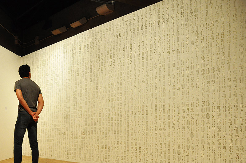 Números da constante pi escritos manualmente em dourado na parede na obra Pi de Felippe Moraes.