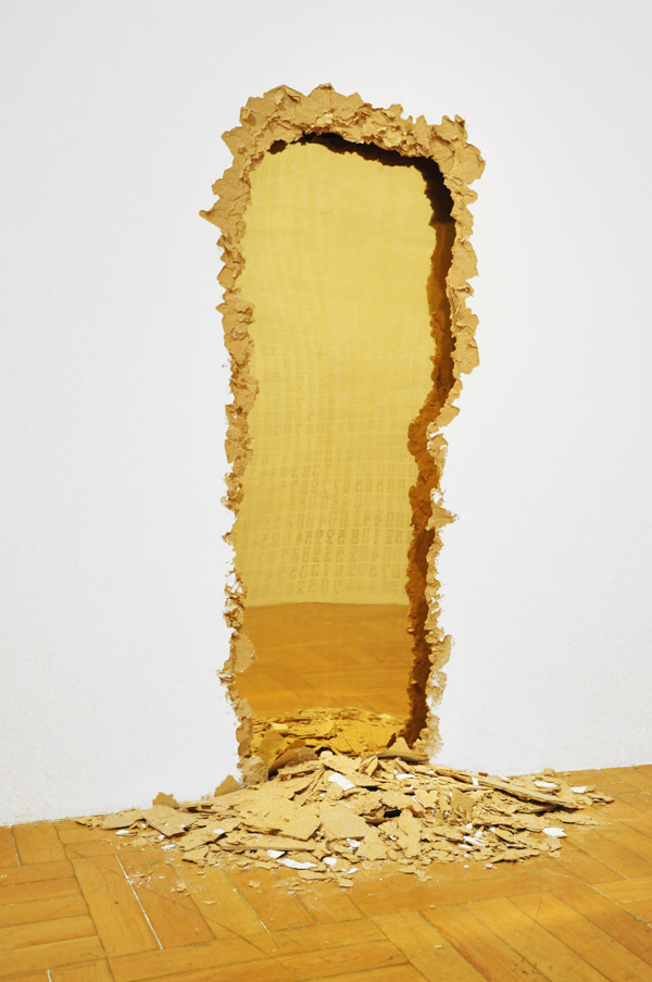 Vista da obra Entranha, intervenção arquitetônica em drywall que revela uma superfície interna de latão polido. A abertura na parede sugere a presença de uma realidade luminosa escondida dentro da arquitetura.