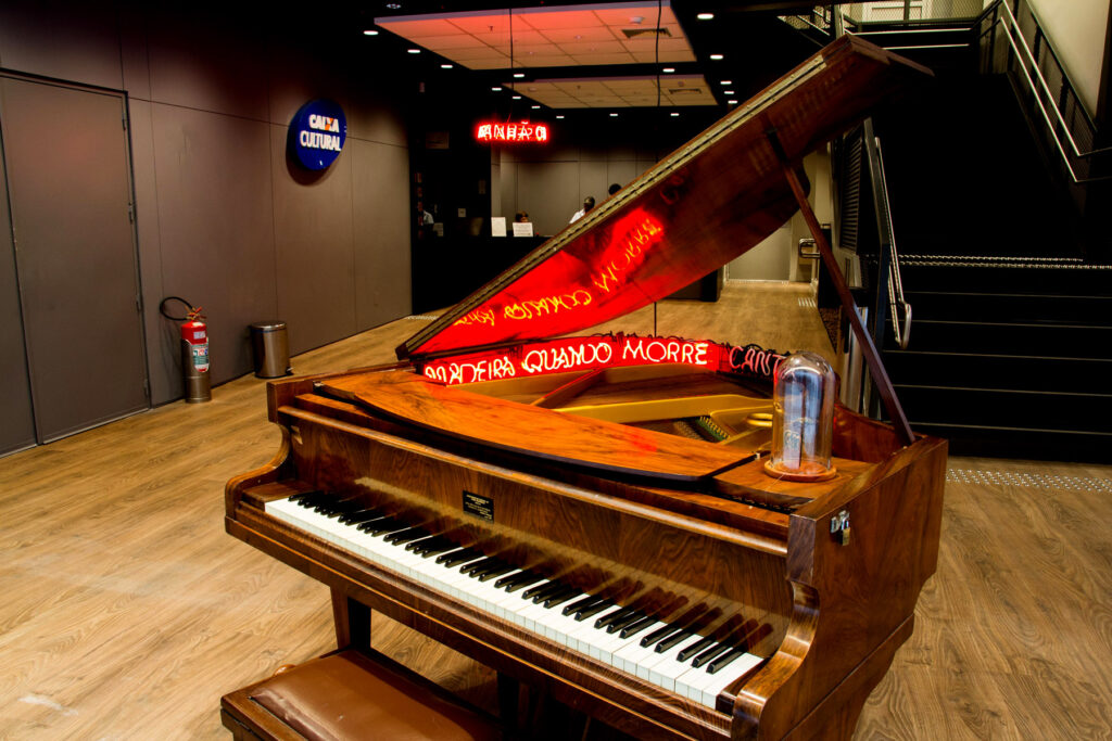 Instalação Madeira quando morre canta de Felippe Moraes com piano de cauda, néon e cigarra sob redoma.