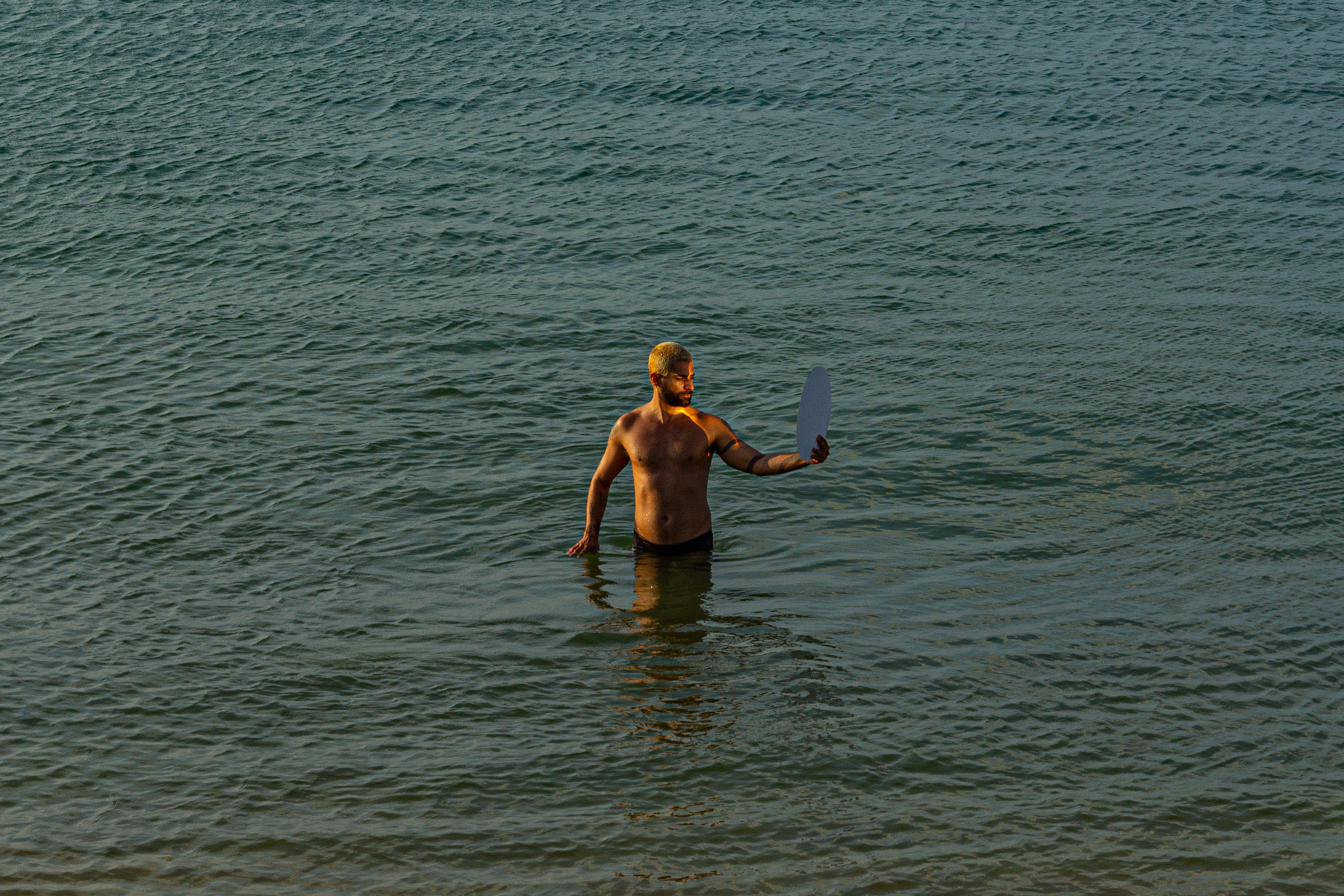 Artista segura objeto circular refletindo luz do sol na praia na obra Narciso acha babado o que é espelho de Felippe Moraes.