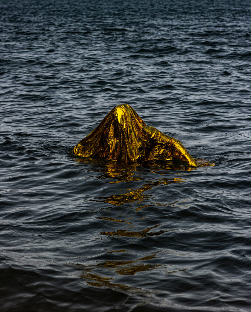 Chamado, obra fotográfica de Felippe Moraes da série Prai'Áurea. Na imagem, uma figura aparece pequena diante da vastidão do oceano segurando uma bandeira dourada que reflete a luz do sol. O gesto sugere comunicação com algo além da linha do horizonte e evoca dimensões simbólicas de busca, sinalização e transcendência.